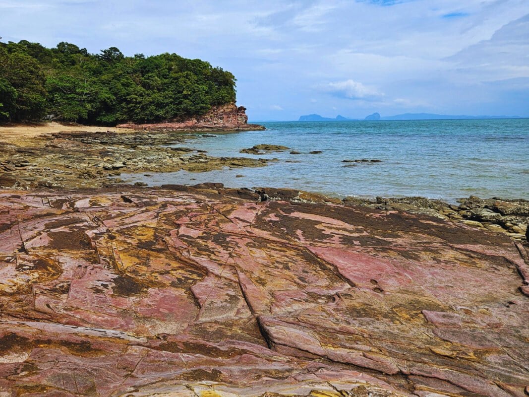 Red rocky shore on Pirate Bay; cliffs covered with jungle and small islands in the distance, Koh Lanta