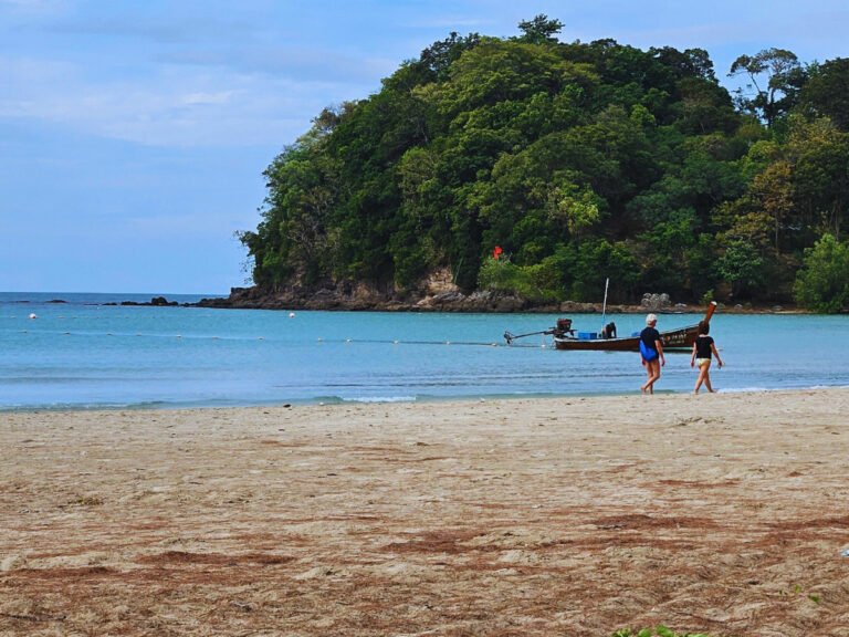 Boat, people walking on the beach, cliff covered with jungle in a distance, Koh Lanta