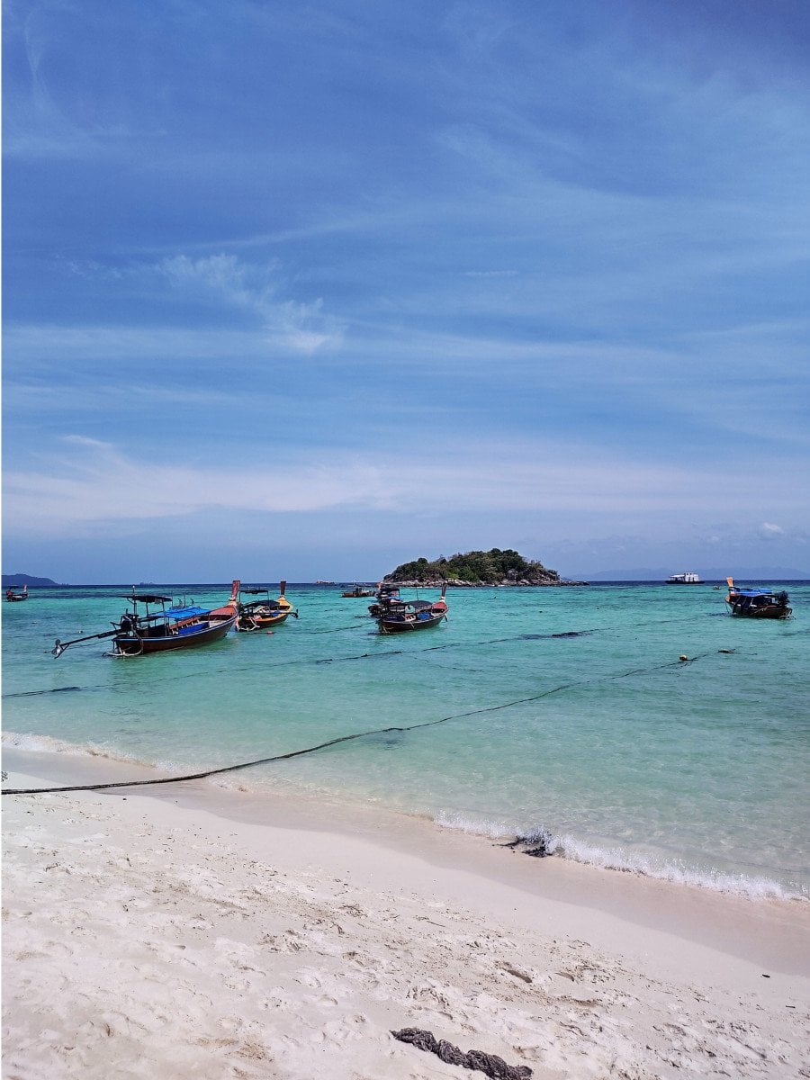 White sandy beach, and boats on Koh Lipe Sunrise beach; small, green islands in the horizon.