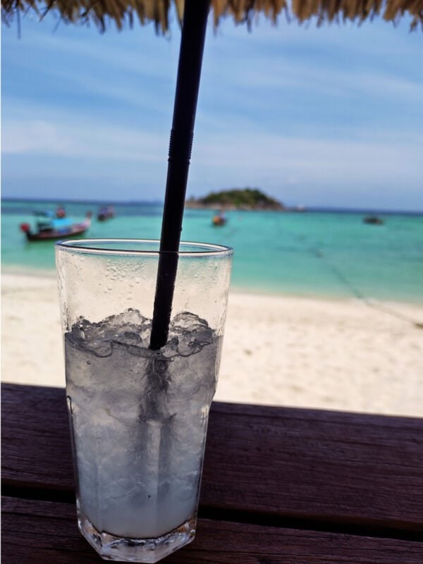 Soda drink in the foreground. White sandy beach, and boats on Koh Lipe Sunrise beach; small, green islands in the horizon.