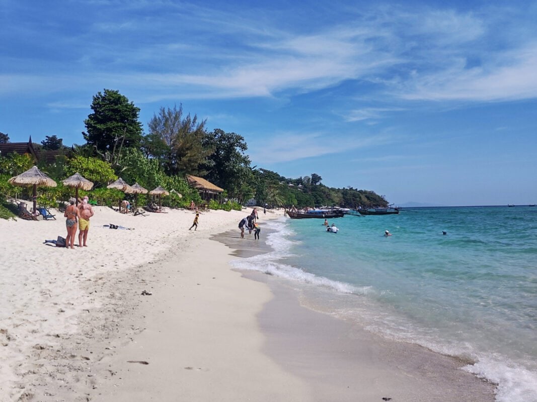 Koh Phi Phi people standing and swimming on a sandy Long beach, boats and trees in the distance