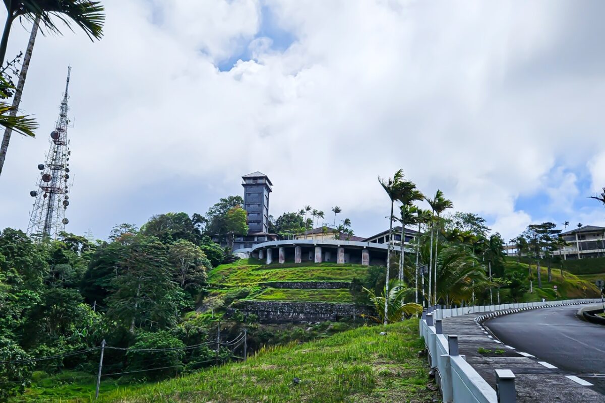 A winding mountain road leads toward a tall observation tower and telecommunications mast under a cloudy sky.