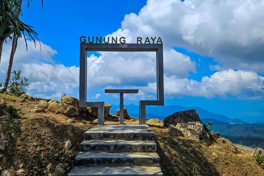 A concrete photo frame monument with "GUNUNG RAYA" text sits on a rocky hill overlooking blue mountains.