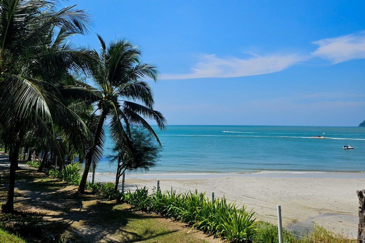 Tropical beach view framed by palm trees with boats on turquoise water under a blue sky on Langkawi.