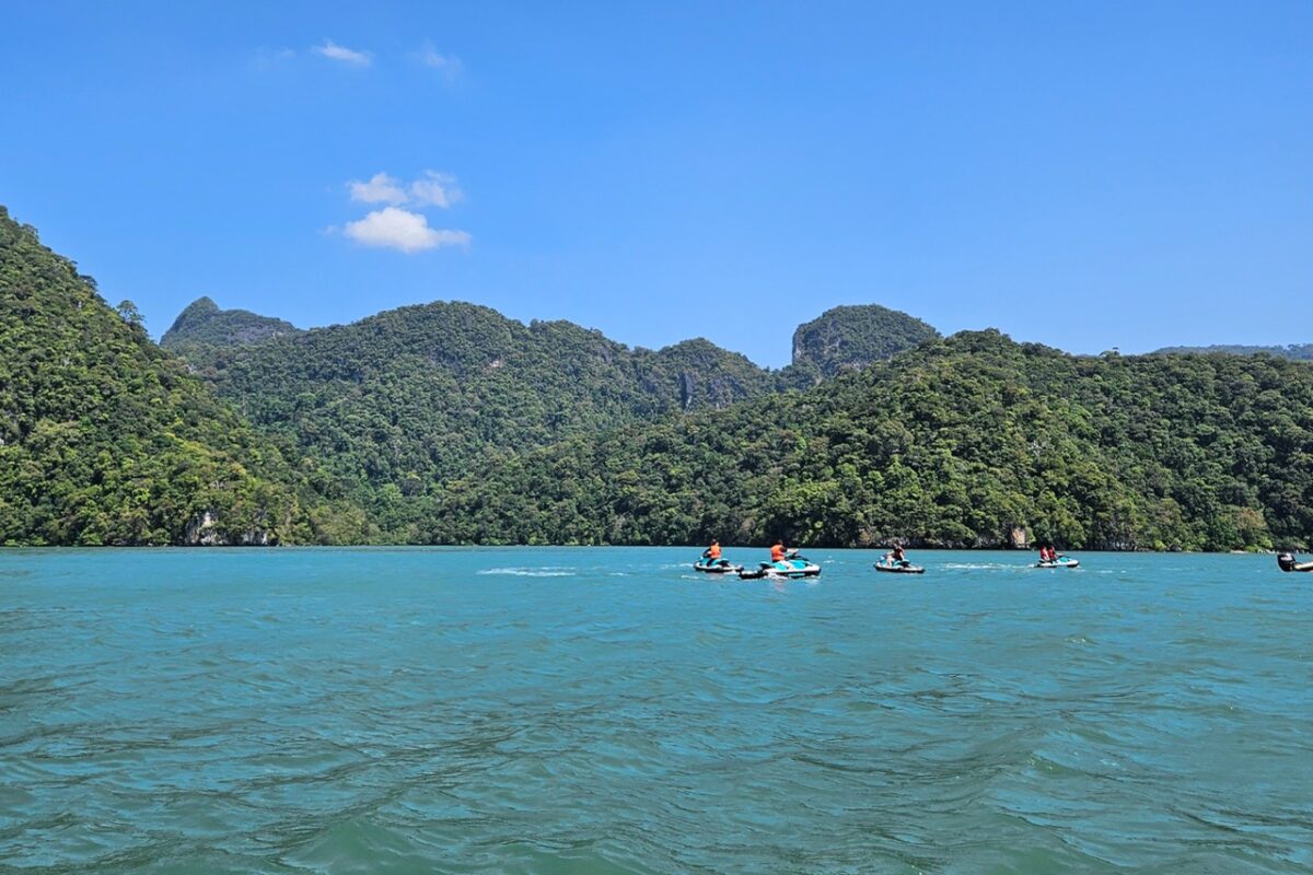 People riding jet skis across bright blue water toward forested island hills, the Pregnant Maiden’s silhouette made of limestone cliffs.