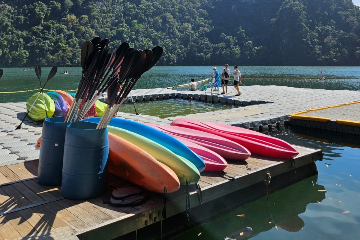 Bright pink, orange, and blue kayaks stacked on a dock next to a bucket of black paddles.