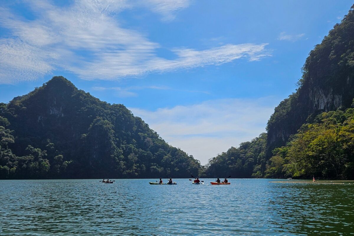 Several colorful kayaks paddling across a wide, calm lake between towering green peaks at Pulau Dayang Bunting Island.